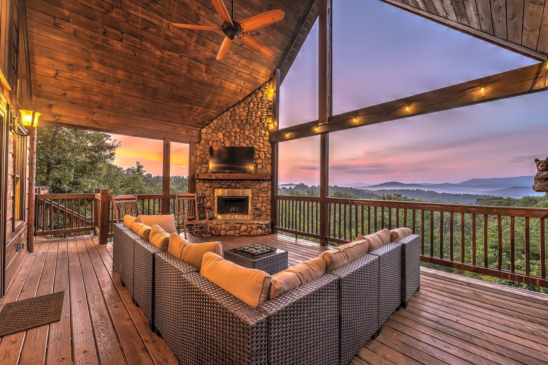 Covered deck at Top of the World in Blue Ridge, GA with a stone fireplace, sectional sofa, and panoramic mountain views at sunset